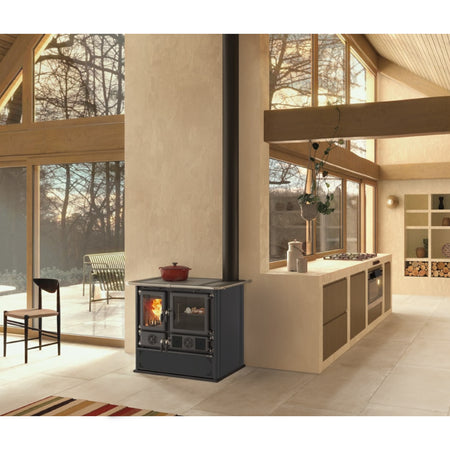 Ambientebild: La Nordica Holzherd Kuechenofen Rosa.16 STEEL, frei im Wohnraum vor grossen Fenstern, Kueche dahinter | Klimaworld.com