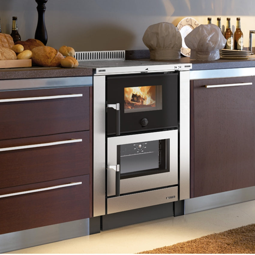 Ambientebild: La Nordica Kuechenofen Vicenza Evo in Edelstahl mit sichtbarem Kaminfeuer und Backfach, integriert in dunkle Kueche | Klimaworld.com